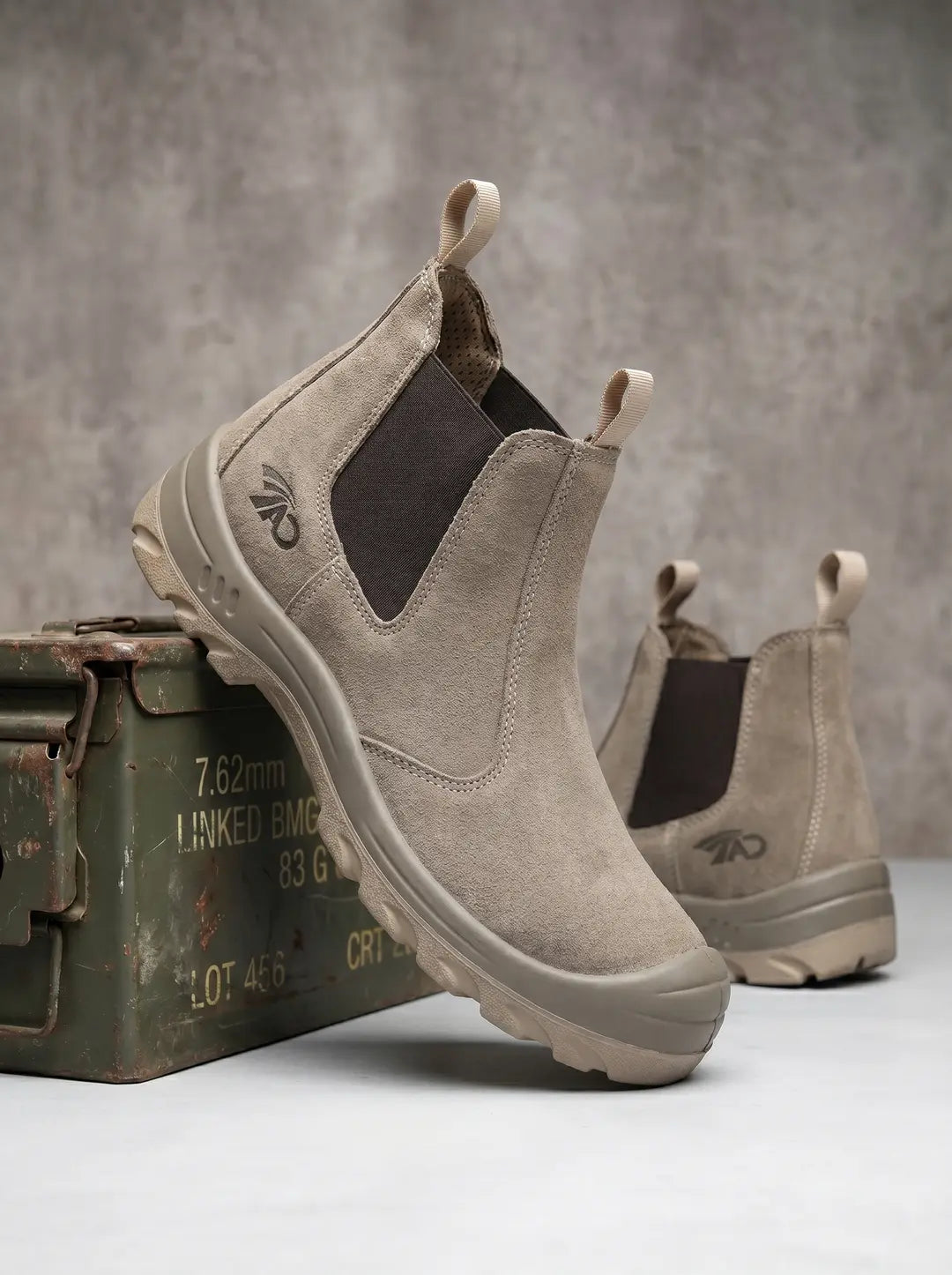 Un par de botas Chelsea de gamuza beige con paneles laterales elásticos marrones y tiradores. Las botas tienen suelas gruesas a juego y un logotipo oscuro en el lateral exterior. Una bota está parcialmente apoyada sobre una caja de municiones militar verde y oxidada, con la segunda bota visible en el fondo.
