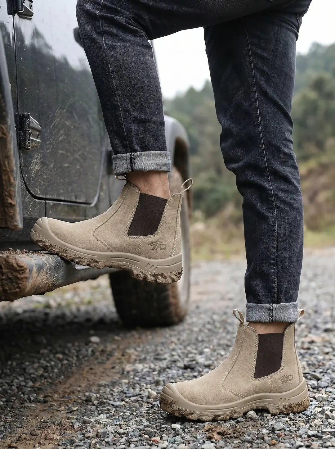 Primer plano de unas botas de trabajo color beige en los pies de una persona. Una bota está sobre el estribo embarrado de un vehículo oscuro y la otra sobre un camino de grava y tierra. Las botas tienen paneles laterales elásticos y suelas robustas y con relieve.