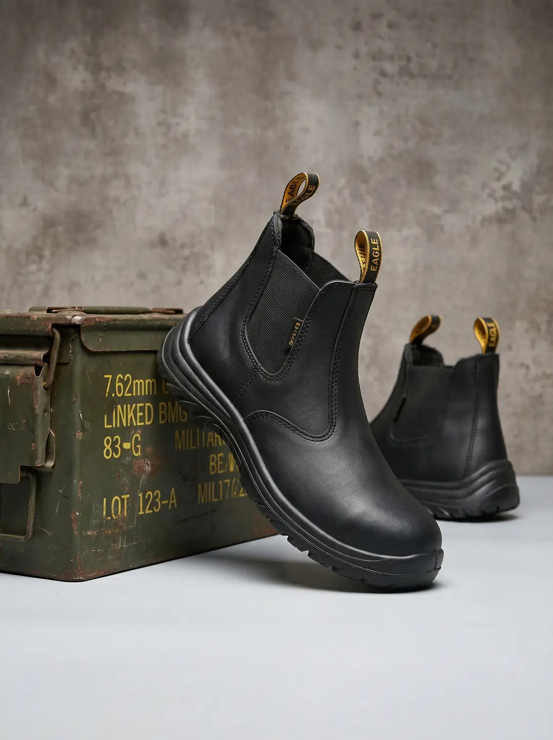 Dos botas de trabajo tipo Chelsea negras, con puntera redondeada, suelas gruesas y paneles elásticos laterales. Ambas botas tienen tiradores amarillos con la inscripción 'EAGLE' en la parte trasera. Una caja de municiones militar verde y oxidada se ve a la izquierda. Fondo gris.