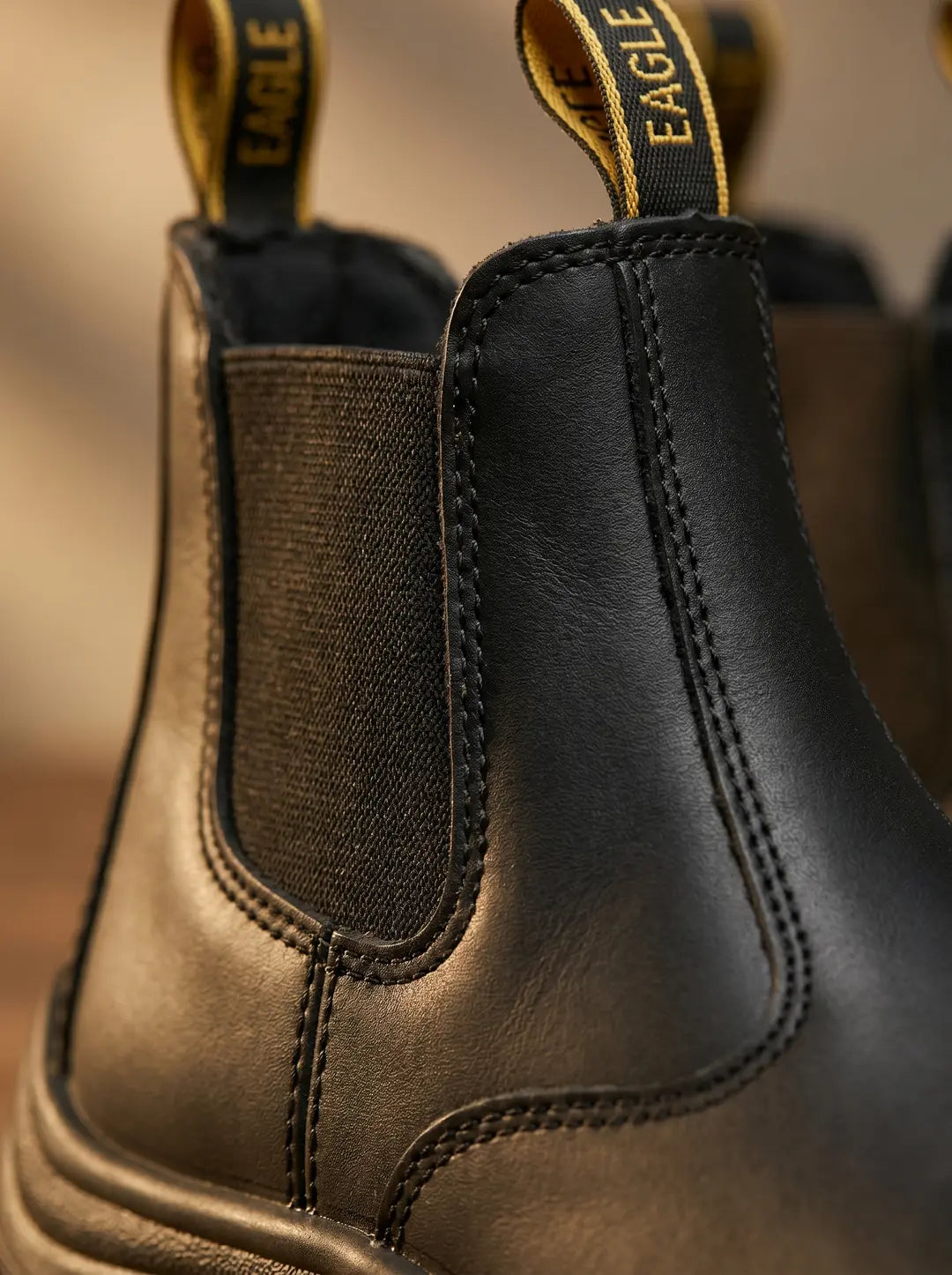Bota negra de cuero con panel lateral elástico negro, costuras visibles, y una tira de agarre trasera amarilla y negra con la palabra 'EAGLE' inscrita. Parte de la suela negra visible en la base.