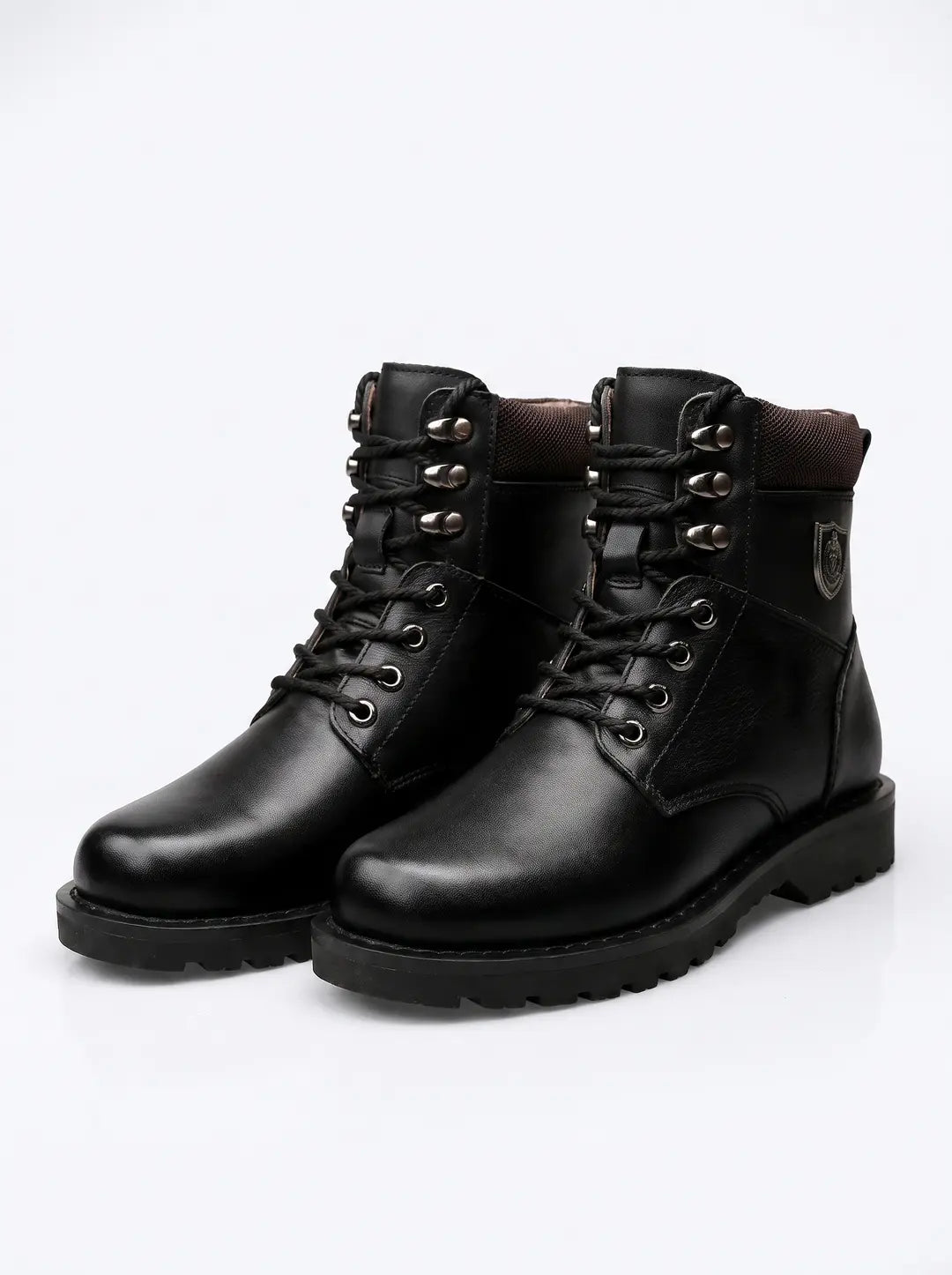 Un par de botas negras con cordones. Presentan ojales y ganchos de metal plateado para los cordones. El cuello de la bota tiene un borde interior de tela texturizada en tono oscuro. Un pequeño emblema metálico adorna el lateral exterior de una de las botas. Las suelas son gruesas y de color negro con relieve.