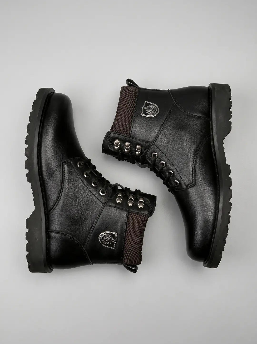 Un par de botas negras de cuero con cordones, ojales y ganchos metálicos plateados. Presentan un cuello superior de tela texturizada en tono marrón oscuro y una insignia metálica plateada en el lateral. Las botas tienen suelas gruesas y están dispuestas sobre un fondo gris claro.