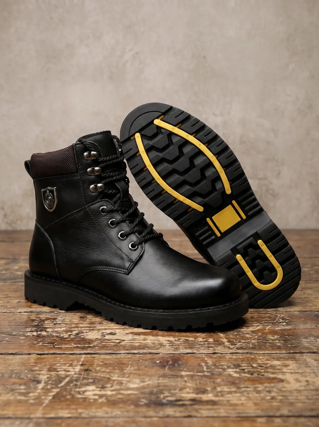 Dos botas negras de cuero. Una bota está de pie con cordones negros, ojales plateados, cuello acolchado marrón y un emblema metálico plateado en el lateral. La otra bota está tumbada, mostrando su suela negra con un patrón de tacos y detalles amarillos brillantes. Ambas están sobre una superficie de madera desgastada con una pared de color claro de fondo.