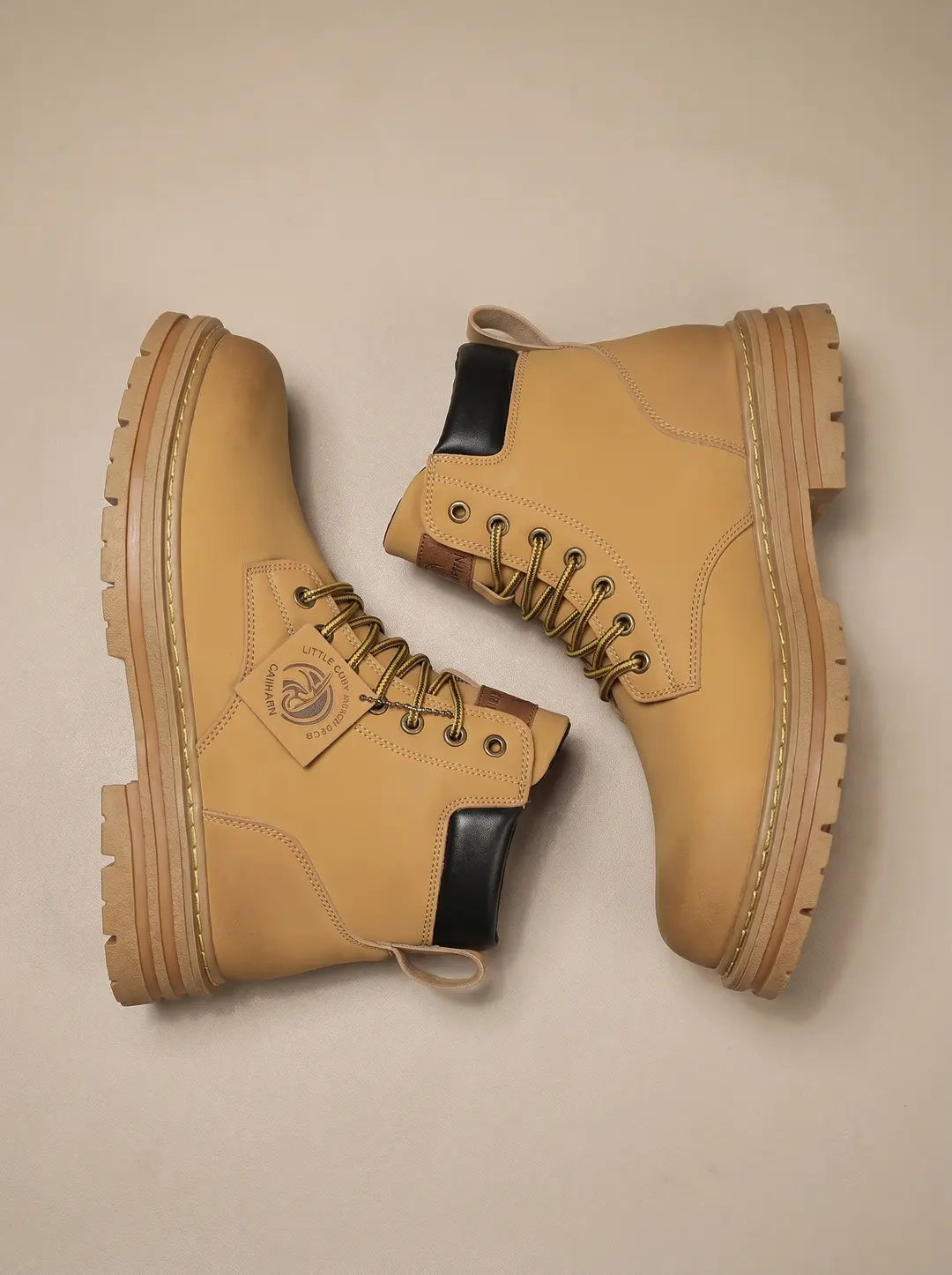Un par de botas de color camel con cordones. Las botas tienen suelas gruesas y texturizadas de color a juego, detalles negros en el área del tobillo y cordones de color dorado. Una etiqueta colgante está unida a una de las botas.