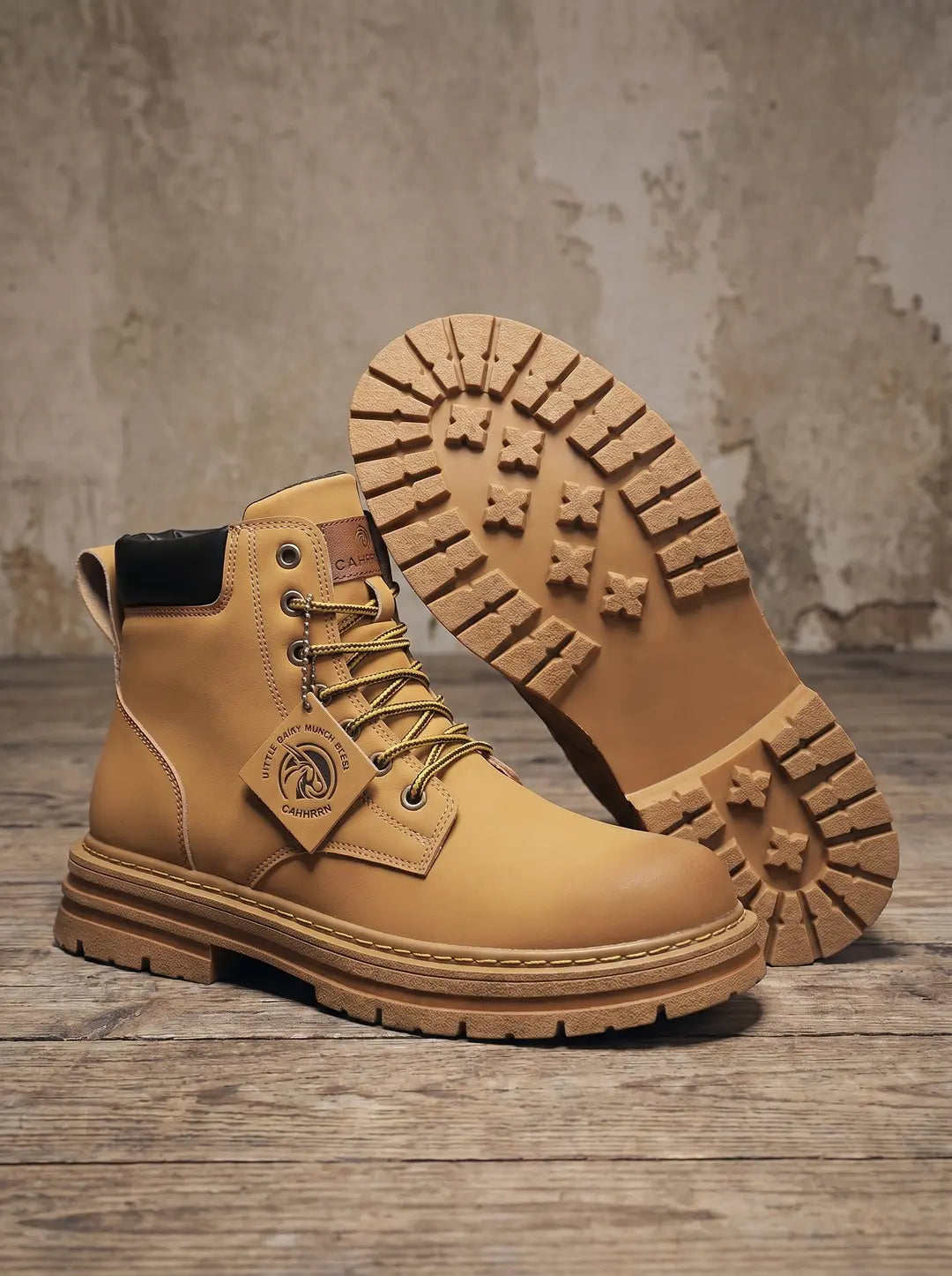 Un par de botas de color camel con cordones amarillos y ojales metálicos. Una bota está de pie mostrando el lateral y la puntera, con un cuello acolchado negro en el tobillo y una pequeña etiqueta colgando de los cordones. La otra bota está tumbada detrás, mostrando la suela gruesa con un patrón de tacos prominente. Las botas están sobre un suelo de madera rústica con una pared texturizada de fondo.