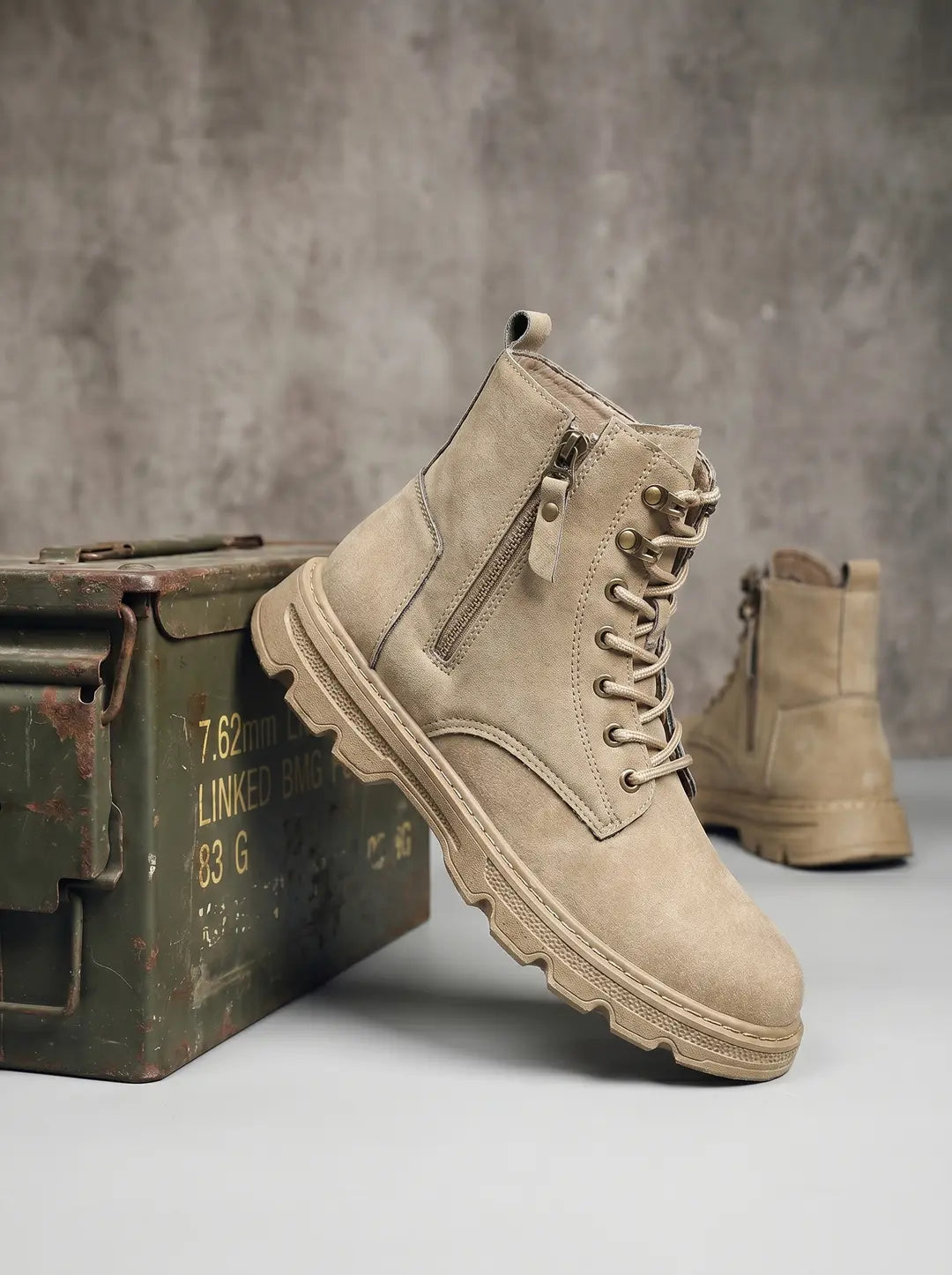Una bota de color beige con cordones, ojales metálicos y una cremallera lateral. Tiene una suela gruesa y dentada. Se apoya en una caja de municiones verde oliva oxidada. Otra bota similar está desenfocada en el fondo.