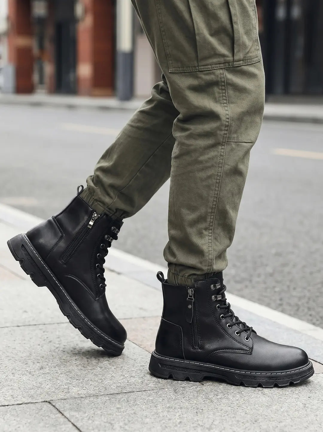 Botas negras de caña alta con cordones y cremallera lateral, con suela gruesa y costuras blancas, usadas con pantalones cargo de color verde oliva en una acera.