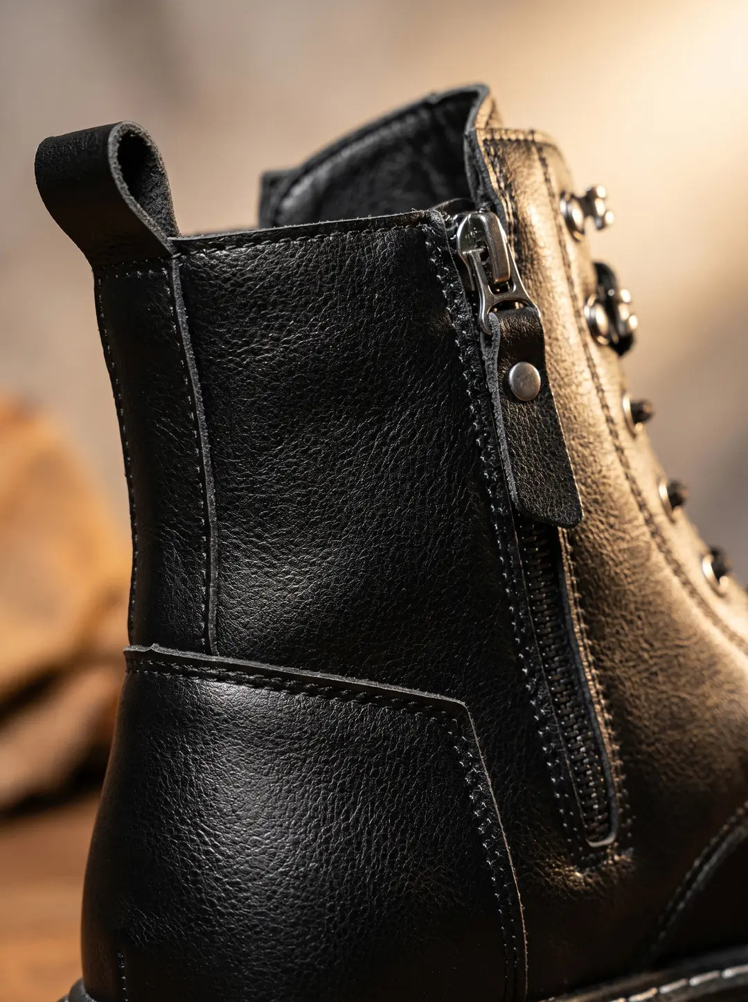 Primer plano de la parte superior y lateral de una bota de cuero negro con textura granulada. Se observa una cremallera lateral metálica, un tirador de cremallera de cuero, una trabilla trasera y detalles de costuras.