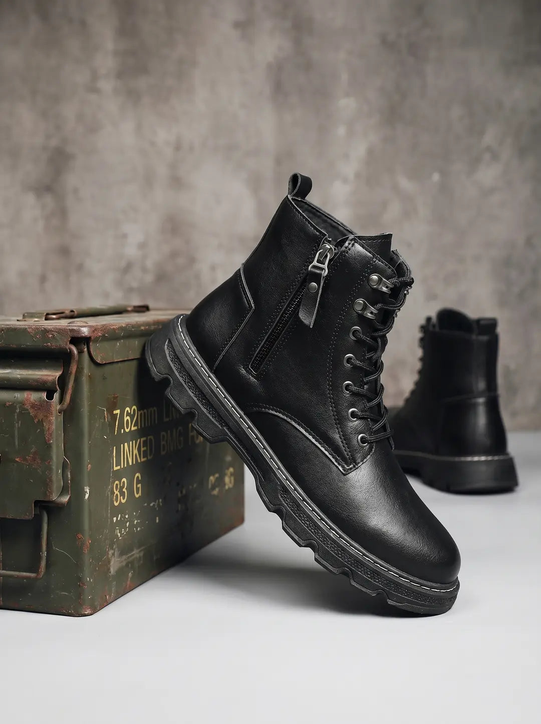 Bota negra con cordones y cremallera lateral, con suela gruesa texturizada y costuras blancas visibles. Se apoya sobre una superficie clara junto a una caja de municiones verde militar oxidada, mientras otra bota similar se ve desenfocada en el fondo.