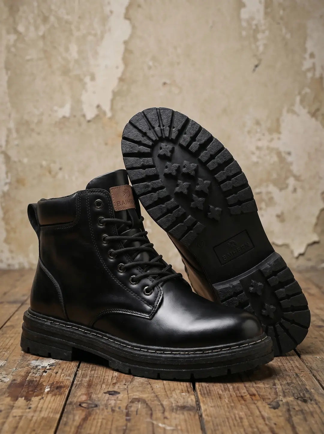 Dos botas negras estilo militar con cordones y suelas gruesas, una en posición vertical y otra de lado mostrando su suela con patrón de tacos. Se observa una etiqueta marrón con la marca BAIHERN en una bota. Las botas están sobre un suelo de madera con una pared clara y texturizada de fondo.
