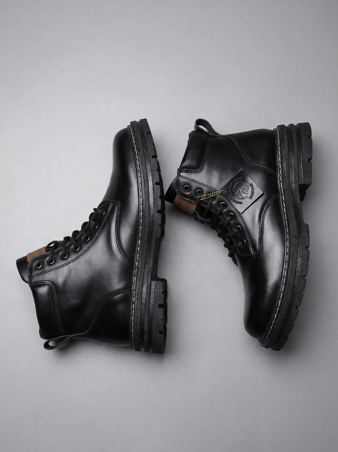 Dos botas de cuero negras con cordones, suelas gruesas y costuras visibles en un fondo gris. Una de las botas tiene una etiqueta colgando.