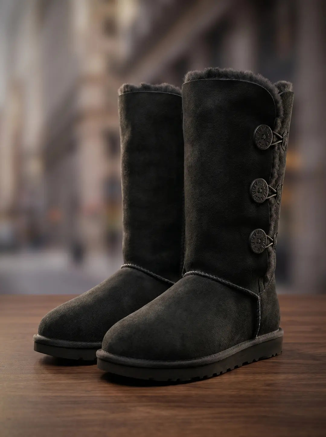 Un par de botas de gamuza de color gris oscuro con forro de piel visible en la parte superior. La bota derecha tiene tres botones circulares oscuros con texto grabado y trabillas elásticas en el lateral exterior. Las botas están sobre una superficie de madera.