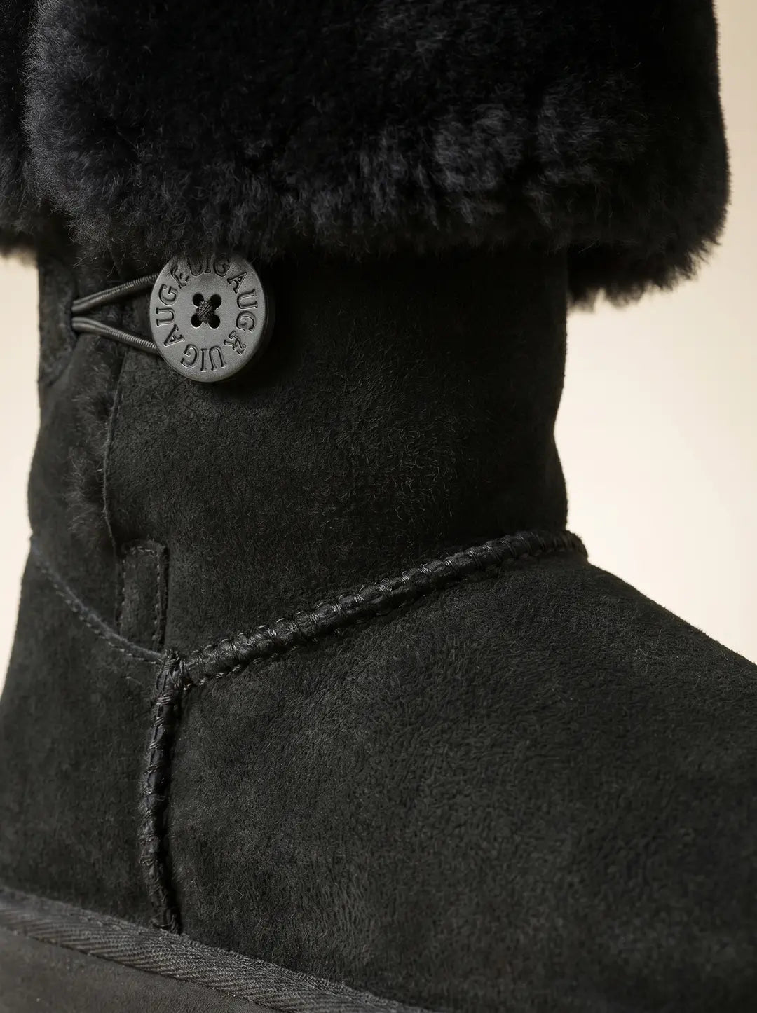 Bota negra de caña corta con ribete de pelo negro en la parte superior y cuerpo de gamuza. Presenta un botón circular negro grabado con la inscripción 'UIGAUG' y una presilla elástica en el lateral. Detalles de costura visible.