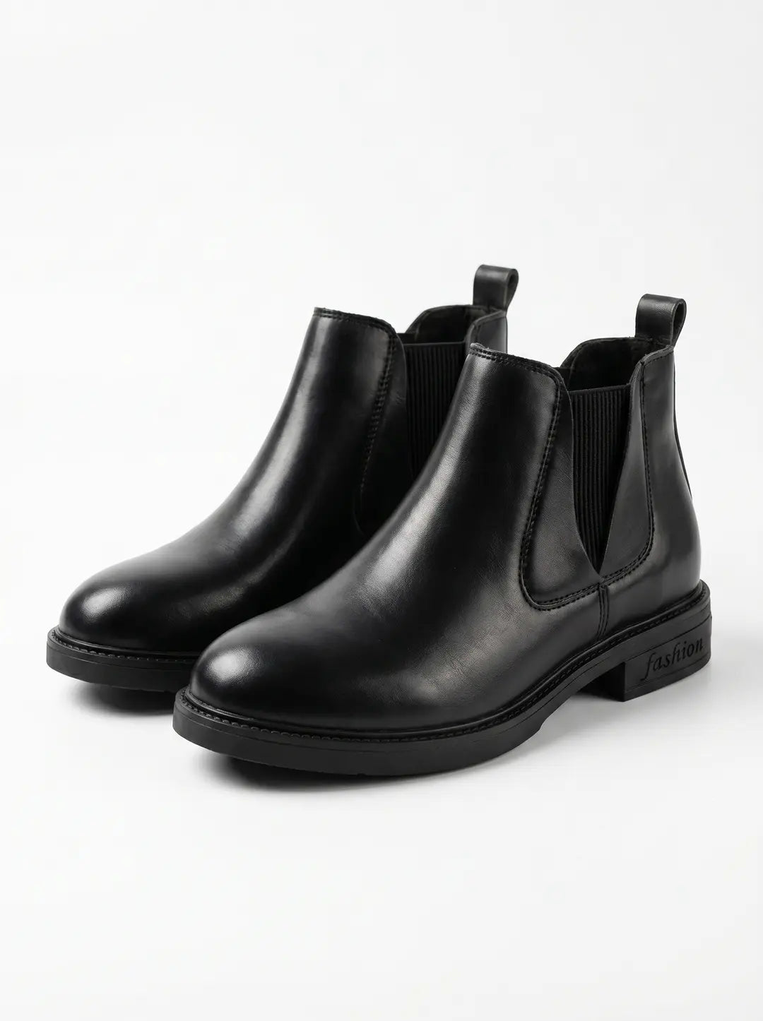 Un par de botines Chelsea negros de cuero con paneles laterales elásticos, tiradores traseros y suelas negras. La palabra 'fashion' está grabada en el tacón de la bota derecha.