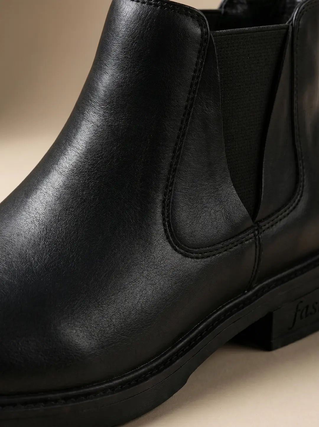 Primer plano de una bota negra con textura similar al cuero, diseño tipo Chelsea con panel elástico lateral y costuras visibles. Suela negra y robusta con la palabra 'fashion' grabada en el talón.