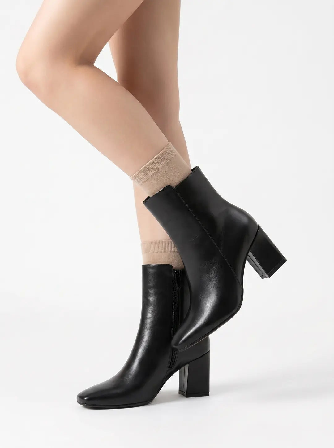 Dos piernas vistiendo calcetines tobilleros de color beige y botines negros con tacón de bloque.