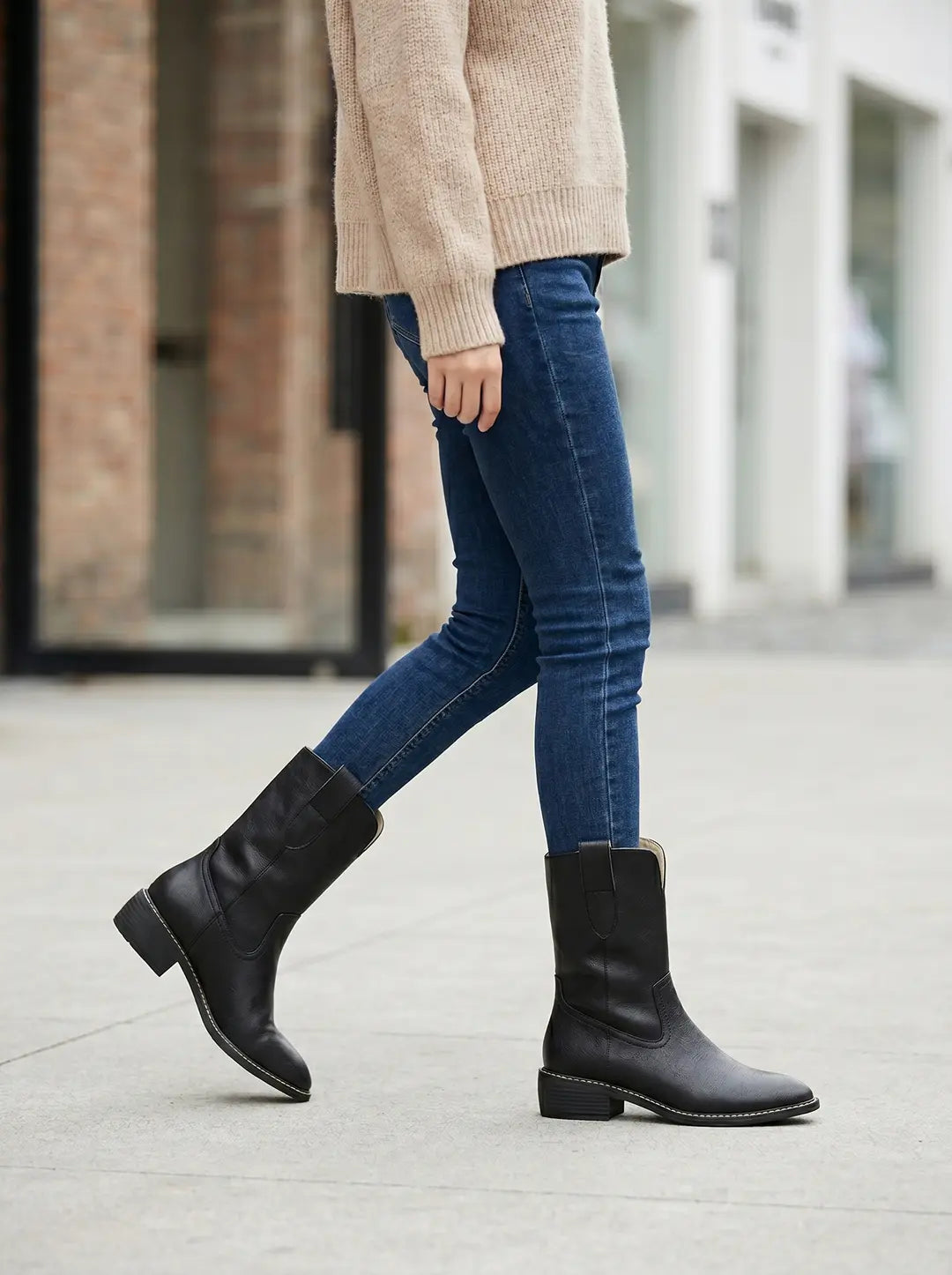Dos botas negras de caña media con tacón bajo y costuras claras visibles a lo largo de la suela, puestas en los pies de una persona que lleva jeans azules y un suéter beige.