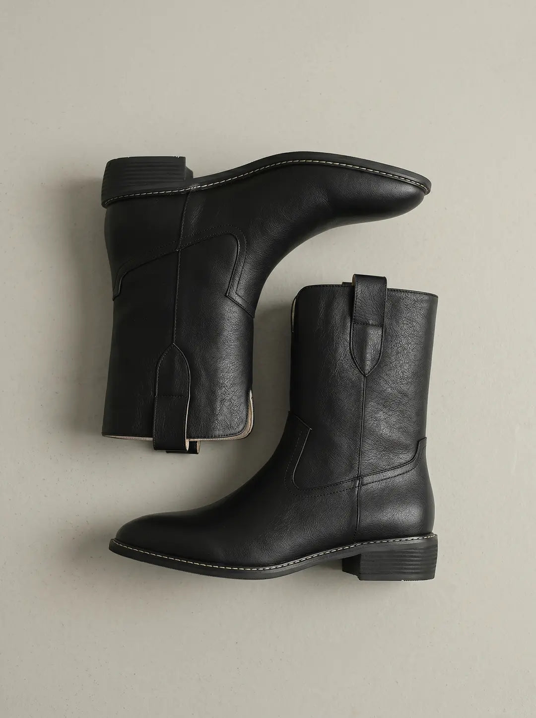 Dos botas negras de estilo vaquero con textura de cuero, exhiben costuras blancas alrededor de la suela y un tacón bajo, dispuestas sobre una superficie gris clara.