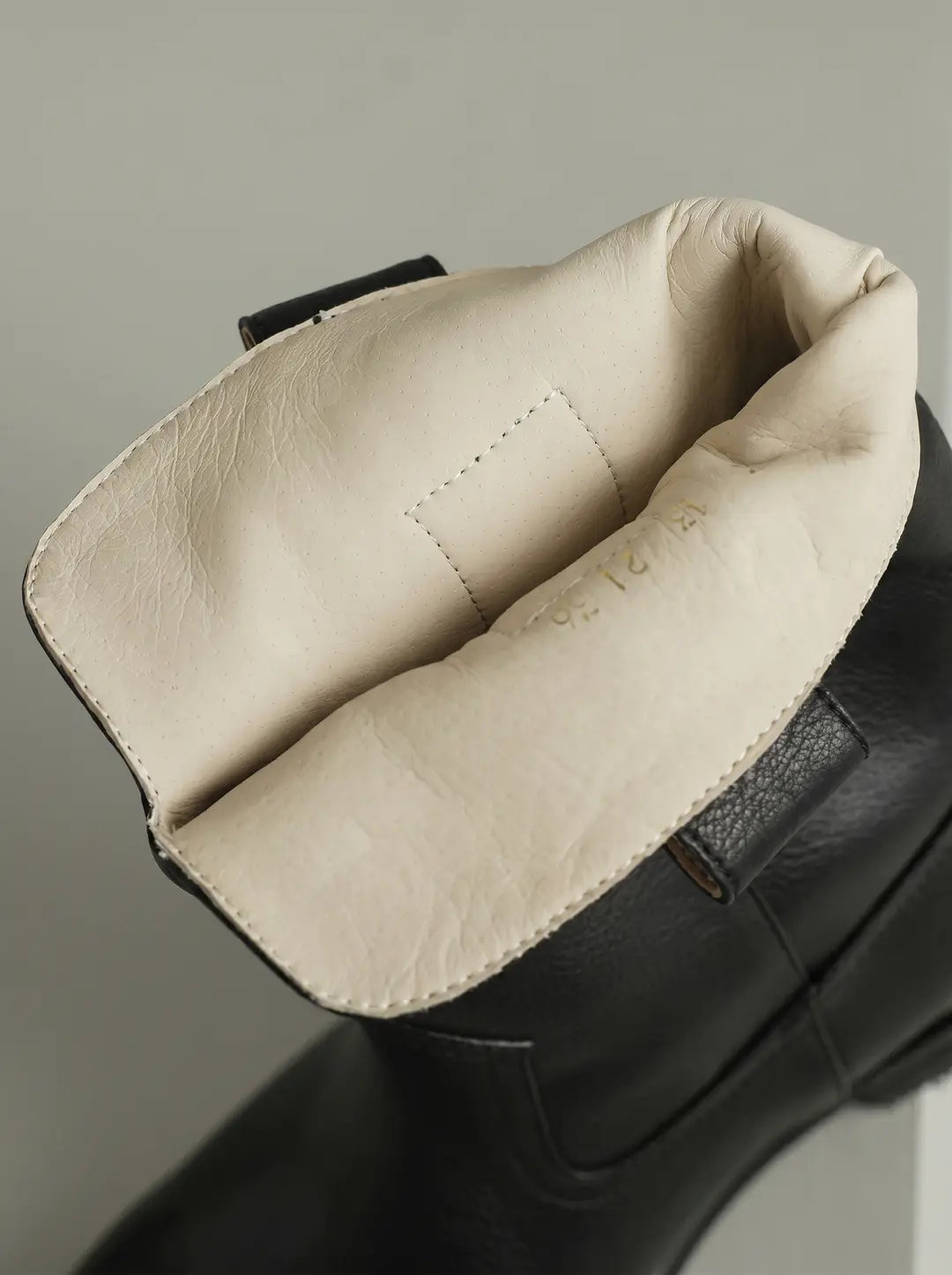 Primer plano del interior de una bota de cuero negra con forro color crema claro. El forro muestra textura de piel con perforaciones sutiles y tiene números dorados impresos. Una pequeña tira de cuero negra se ve en el borde superior.