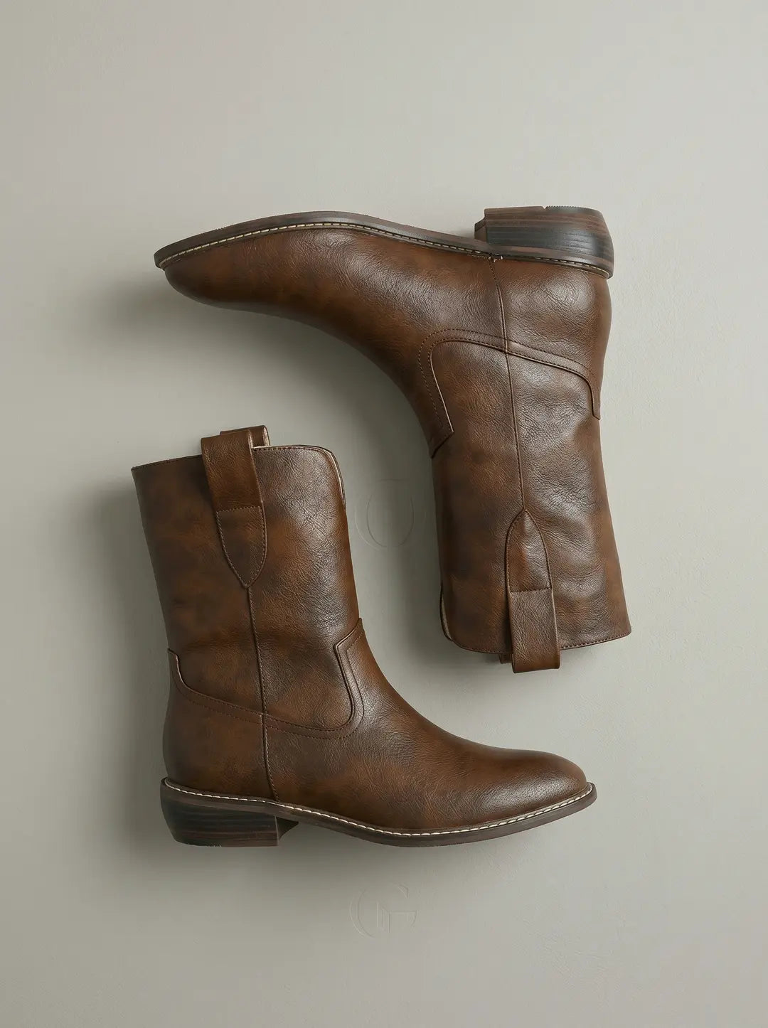 Par de botas marrones de caña media con textura de cuero sintético, costuras decorativas claras en la suela y tacones bajos apilados, dispuestas sobre un fondo liso de color gris claro.